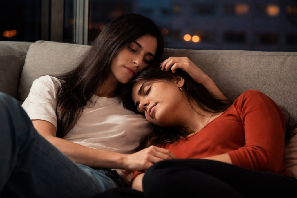 Duas pessoas sentadas em um sofá à noite, uma delas com a cabeça apoiada no ombro da outra, em gesto silencioso de cuidado e proximidade, simbolizando como atitudes simples e cotidianas, quase imperceptíveis, vão definindo aos poucos o rumo emocional de um vínculo.