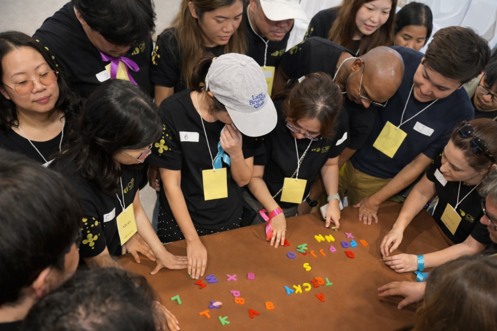 Grupo diverso de pessoas reunidas em atividade colaborativa ao redor de uma mesa, interagindo e organizando letras coloridas, simbolizando cooperação e construção coletiva de sentido.
